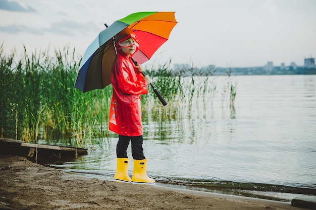 Boy in a red raincoat and yellow rubber boots stands at river
bank and holding rainbow umbrella