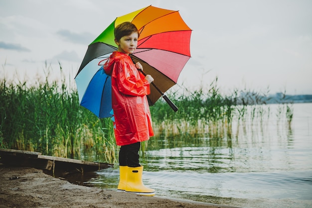 Boy in a red raincoat and yellow rubber boots stands at river bank and holding rainbow umbrella