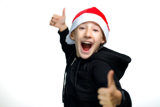 A boy in a red Santa hat who stands on a white background gives a thumbs up and laughs loudly