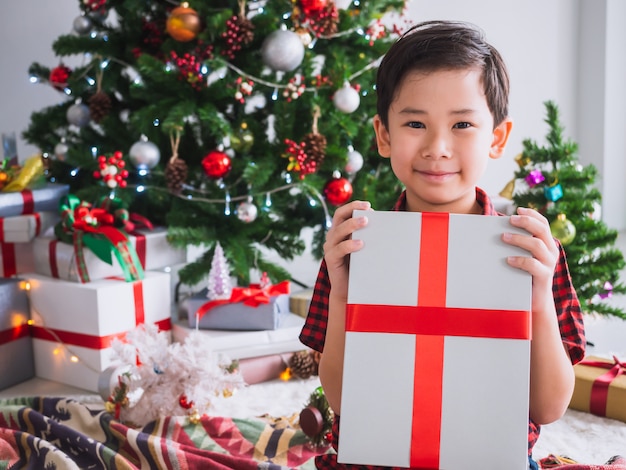 Foto il ragazzo in una camicia rossa sta tenendo il contenitore di regalo ed è felice di celebrare il natale con l'albero di natale