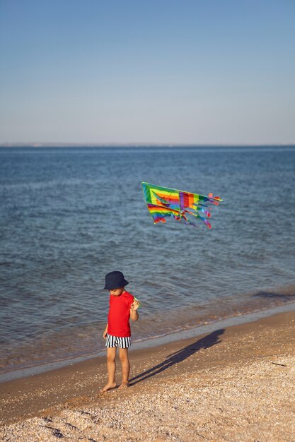 Boy in a red T-shirt and striped swimming trunks launches kite on the beach by the sea in the summer on vacation