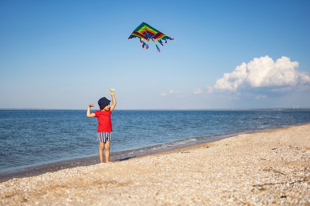 Boy in a red T-shirt and striped swimming trunks launches a kite on the beach by the sea in the summer on vacation