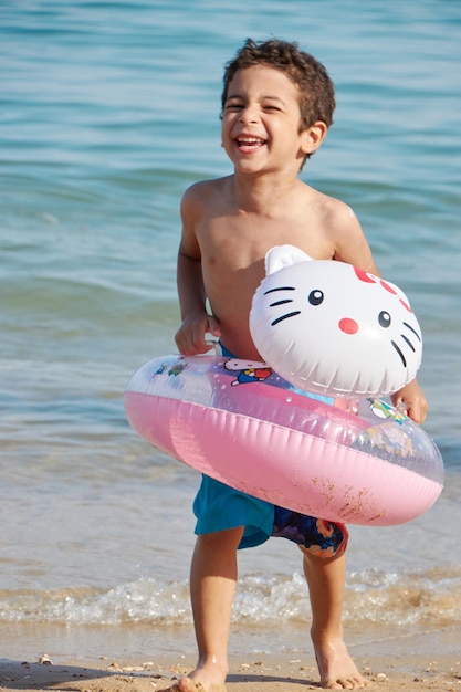 A boy runs on the beach with a hello kitty in his swimsuit.