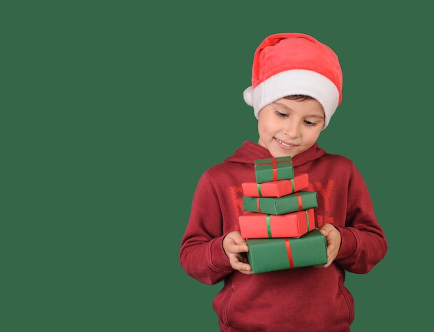 Boy in santa claus hat holding many gifts on green background