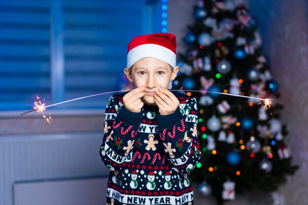 a boy in a Santa Claus hat with sparklers in his hands on the background of a Christmas tree with neon lightin