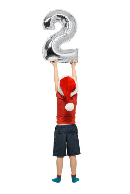 Boy in Santa hat holds up silver inflatable digit two. Back view. Isolated on white background.