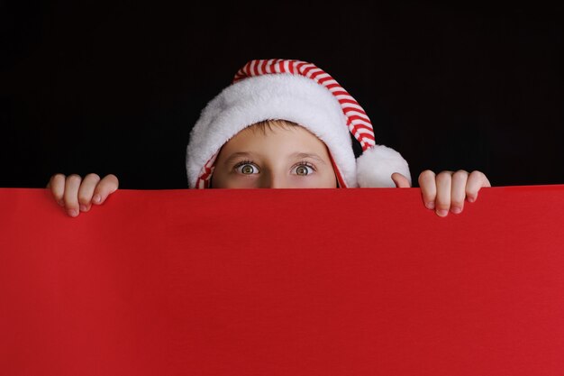 Boy in Santa's hat holding the red blank banner for christmas greetings. Isolated on black