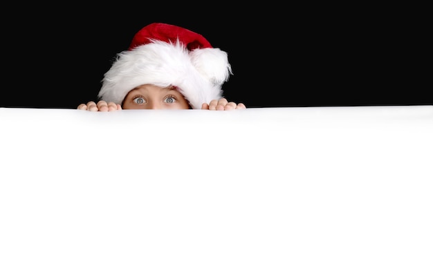 Boy in Santa's hat holding the white blank billboard for christmas greetings. Isolated on black