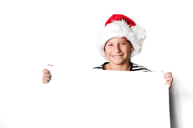 Boy in Santa's hat smiling and holding white blank board for greetings. Isolated on white