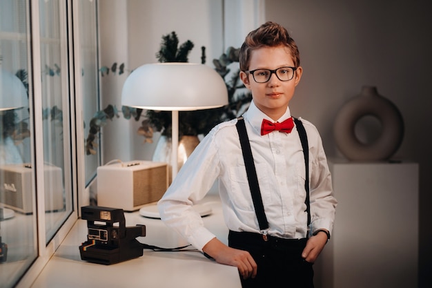 A boy in a shirt with a red bow tie and glasses stands at the window.