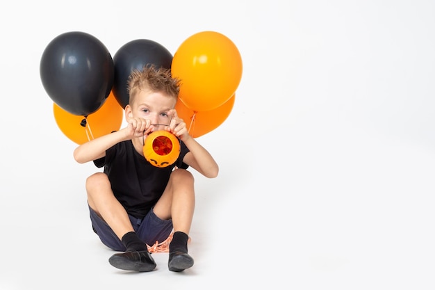 A boy showing an empty bucket in the shape of a pumpkin and holding black and orange balloons eating candy