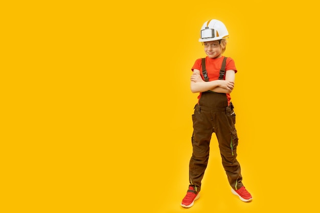 Boy simulates worker wearing helmet and overalls Portrait of child as builder on yellow background Copy space mock up