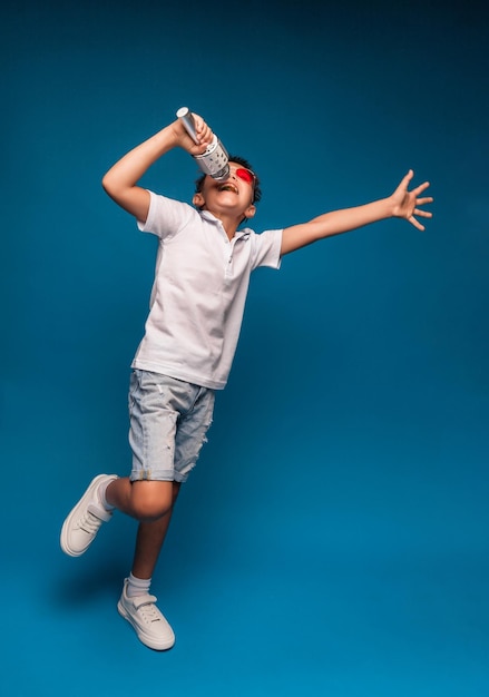 A boy sings into a microphone on a blue background A handsome boy in a white Tshirt and shorts stands on a blue background and emotionally sings into the microphone