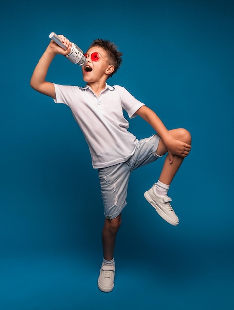 A boy sings into a microphone on a blue background A handsome boy in a white Tshirt and shorts stands on a blue background and emotionally sings into the microphone