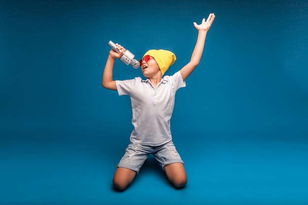 A boy sings into a microphone on a blue background A handsome boy in a white Tshirt and shorts stands on a blue background and emotionally sings into the microphone