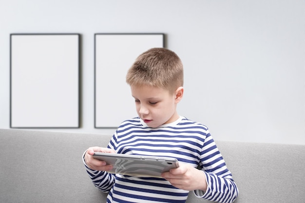 The boy sits on the couch and looks at the tablet