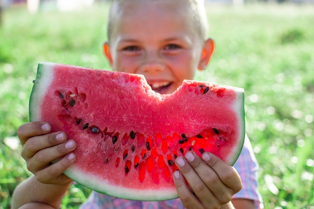 Boy sits on the green grass in the village and holds large piece of watermelon