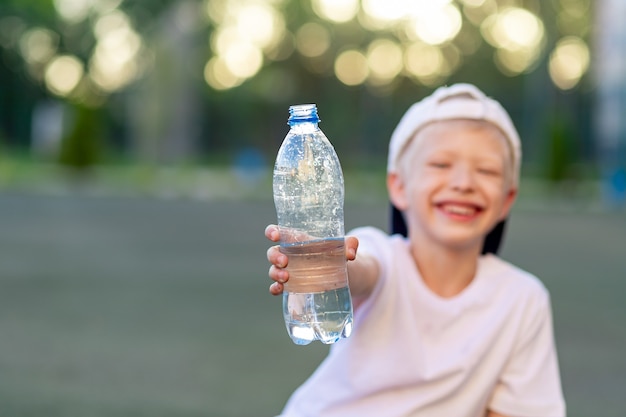 少年はサッカー場の緑の芝生に座って、水のボトルを保持しています。水のボトルに焦点を当てる