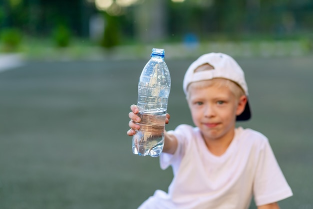 少年はサッカー場の緑の芝生に座って、水のボトルを持っています。水のボトルに焦点を当てます。