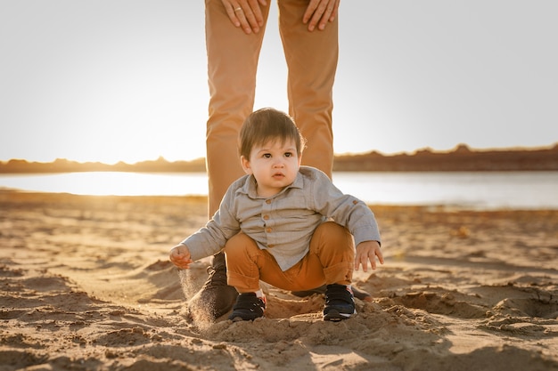 boy sits on his father's feet in the sand on the riverbank.