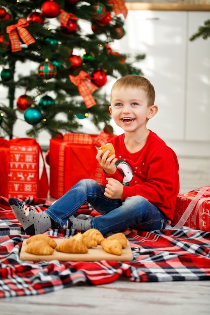 Il ragazzo si siede vicino all'albero di natale nella cucina di natale. tiene un croissant nelle sue mani. sere d'inverno accoglienti a casa. ci sono tanti regali di natale sotto l'albero