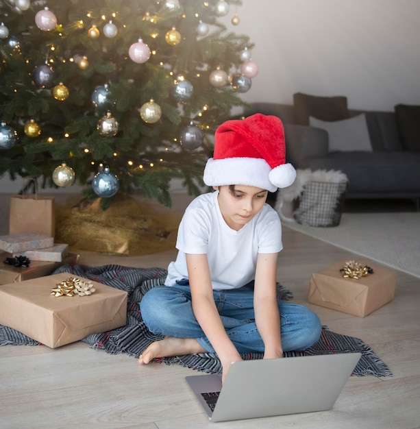 Un ragazzo si siede vicino a un albero di natale con un laptop. albero di natale in un ampio soggiorno.