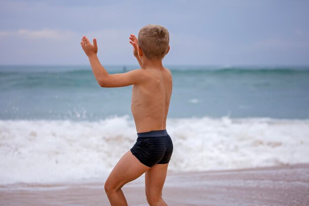 Boy sits on the ocean shore with his arms open towards wind and waves storm in summer courage