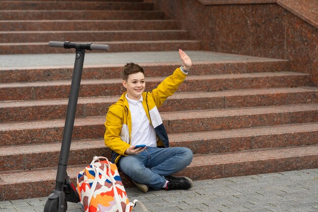 Boy sits on the steps near electric scooter and waving