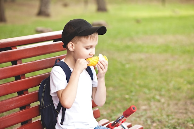 Boy sitting on a bench outdoor in a park and eating fresh boiled corn.