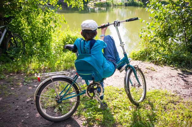 Boy sitting on bicycle on waterside of river among green trees illuminated by the sun