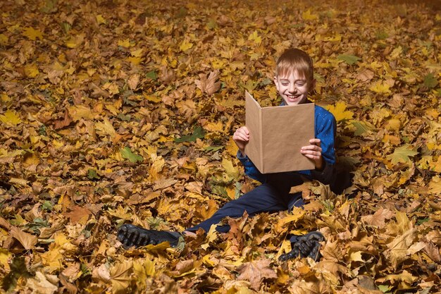 落ち葉の上に座っている男の子は本を読んで笑う 秋の森で本を持つ子供