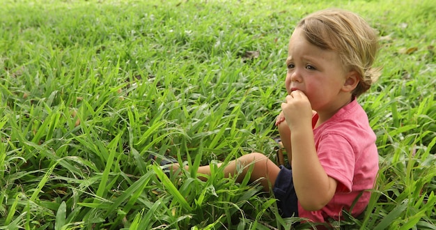 Boy sitting in the grass outside. He is wearing a red shirt side view