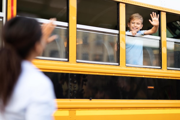 Ragazzo seduto all'interno di un autobus scolastico giallo e che agita la mano alla madre