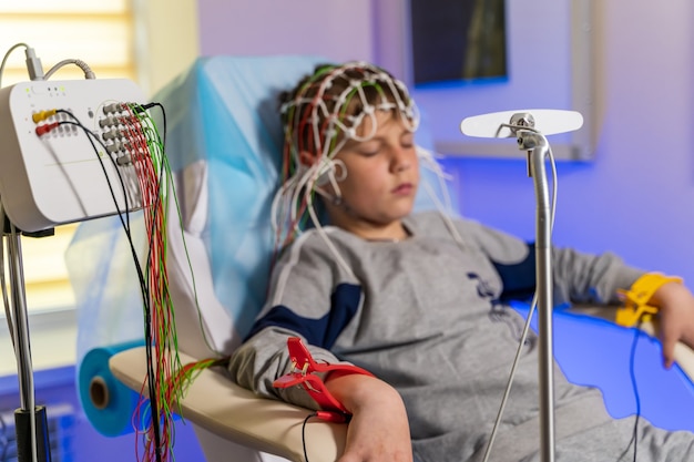 Boy sitting in medical chair. Wearing a special cap during electroencephalography. Monitor with results.