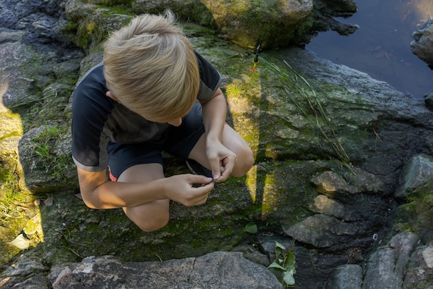 A boy sitting on rocks by the river equips the hook with bait Sport fishing on the river in summer