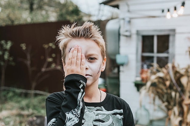 A boy in a skeleton costume with a painted face on the porch of a house decorated to celebrate a Halloween party