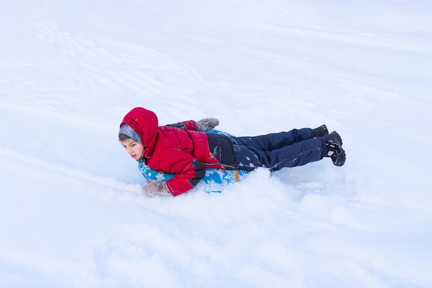 Boy sliding on blue tubing lying on stomach in winter clothes among white snow