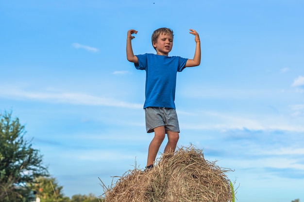 Boy smile play dance grimace show off blue tshirt stand on haystack bales of dry grass clear sky sunny day Balance training Concept happy childhood children outdoors clean air close to nature