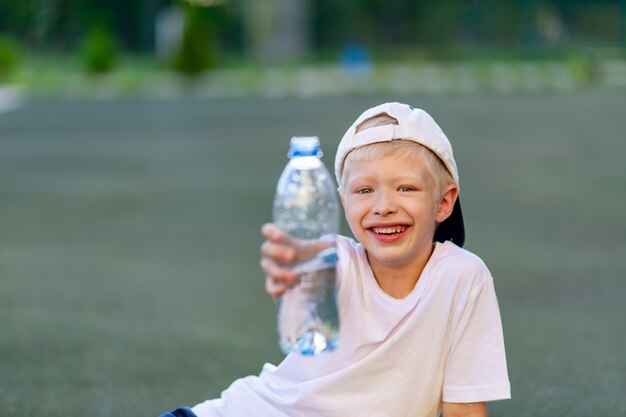 Boy in a sports uniform sitting on a green lawn on a football field and holding a bottle of water