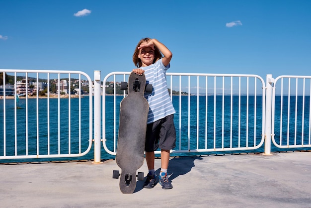 写真 晴れた日に桟橋でロングボードを持って立っている少年