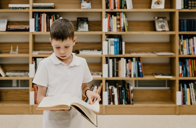 A boy stands in the library and reads a book while standing. Preparing for homework. The boy loves to read. Free space at the school. Extracurricular learning.