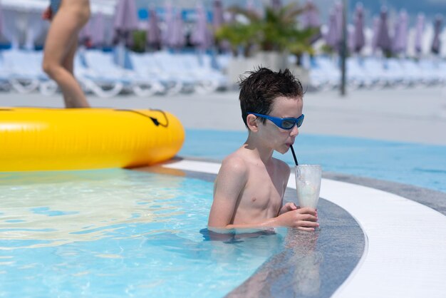 Photo boy in sunglasses is standing in a pool with blue water and drinking a milkshake resort summer