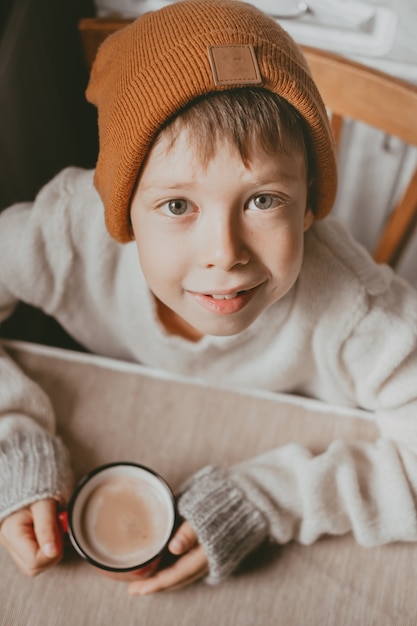 스웨터와 갈색 모자를 쓴 소년이 빨간 컵에서 코코아를 마신다. 머그컵을 들고 포근한 사진. 큰 눈을 가진 소년은 아래에서 보이는 상위 뷰