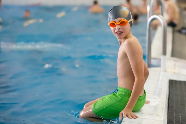 수영 모자와 고글을 쓴 소년이 스포츠 수영장 옆에 앉아 있다