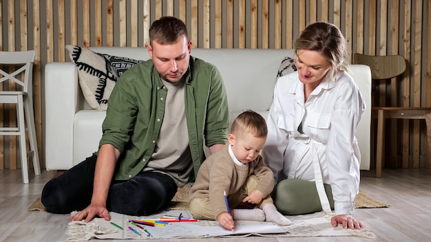 Photo boy takes pencil and draws sitting near parents on floor mat