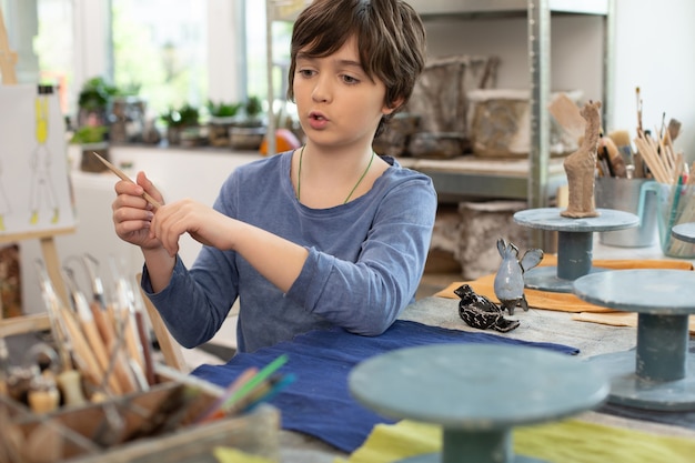 Boy taking little brush while sculpting clay figures
