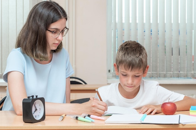 テーブルでノートにテキストを書く宿題をしている少年と教師 母親が息子がノートに書くのを手伝っている