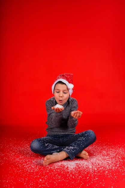 Il bambino ragazzo soffia la neve artificiale dalla sua mano su uno sfondo rosso