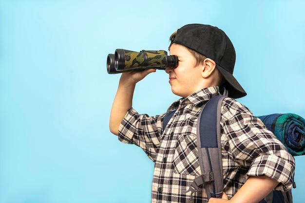 Boy tourist in plaid shirt and cap looks through binoculars on a blue background copy space