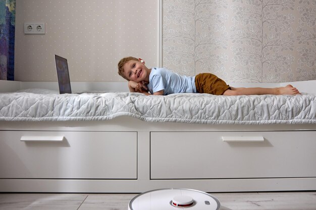 Boy uses a laptop on the bed while the robot vacuum cleaner does the cleaning